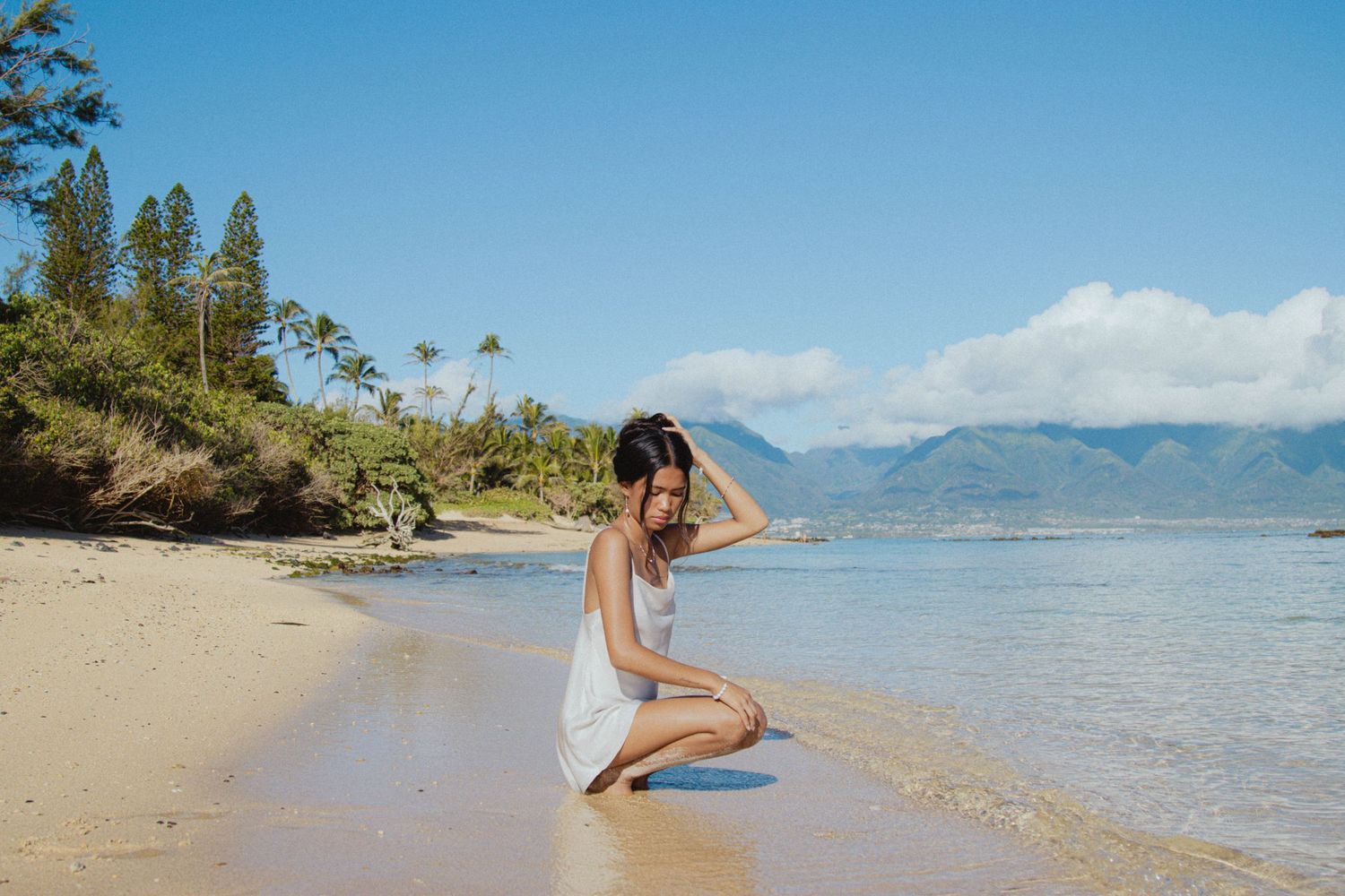 Woman in white dress sitting on a sandy tropical beach with clear blue water and mountains.