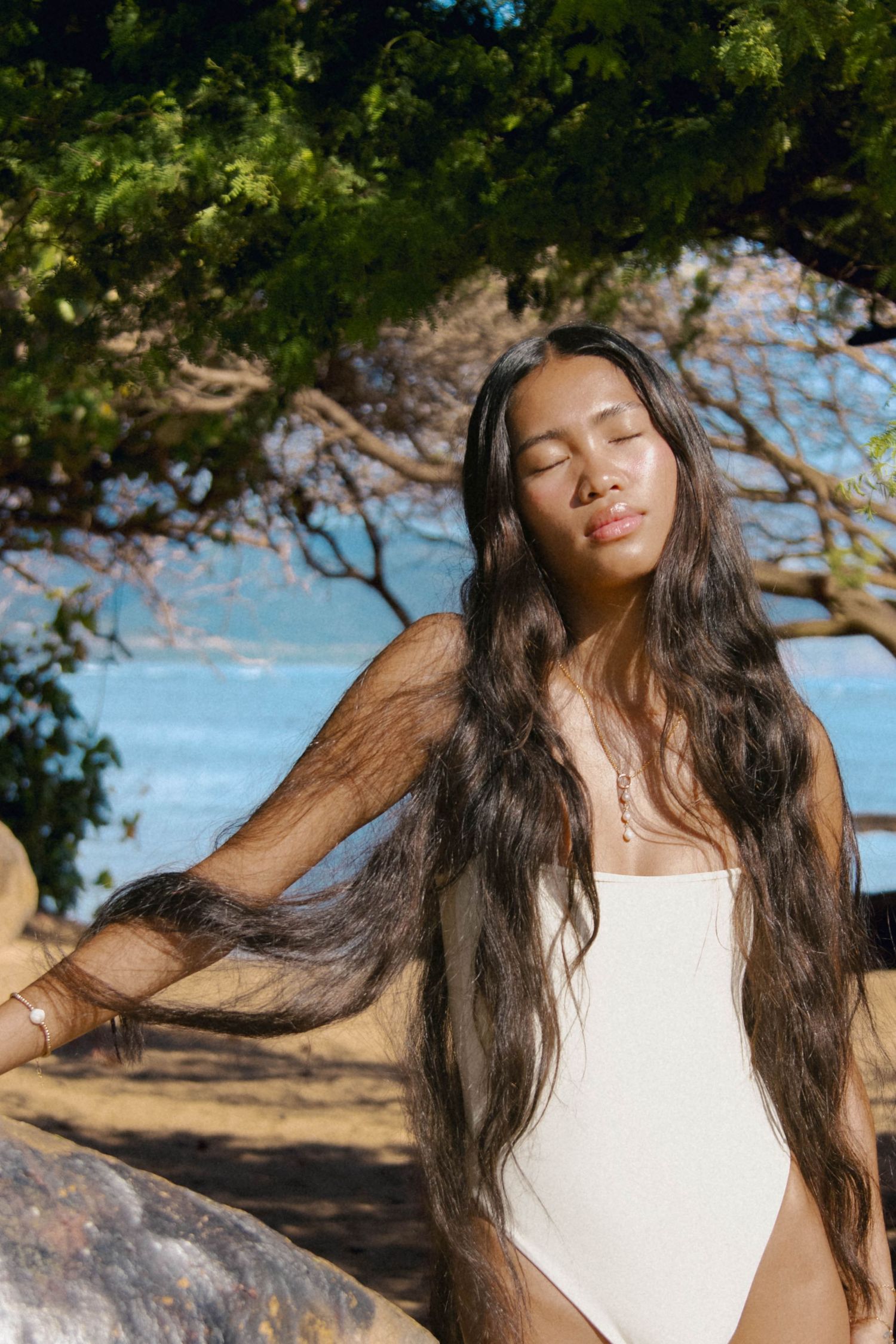 Woman with long dark hair and closed eyes in white swimsuit posing near ocean with trees behind.