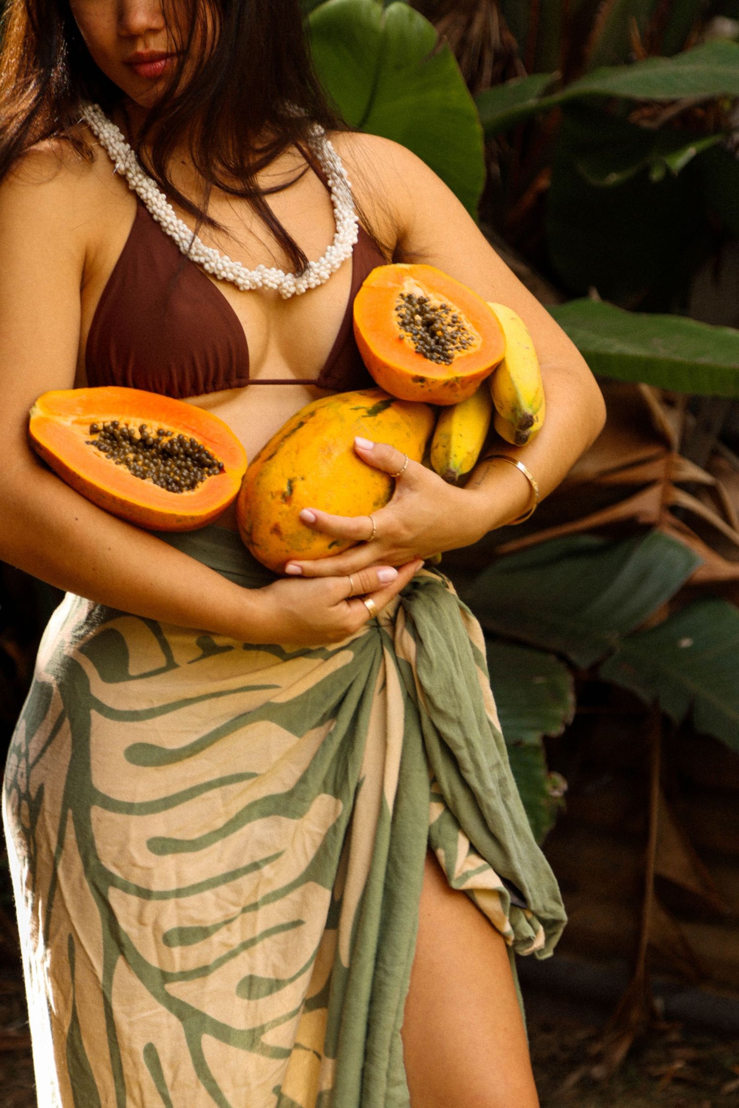 Woman in brown bikini top and sarong holds fresh papayas and bananas in tropical garden setting.