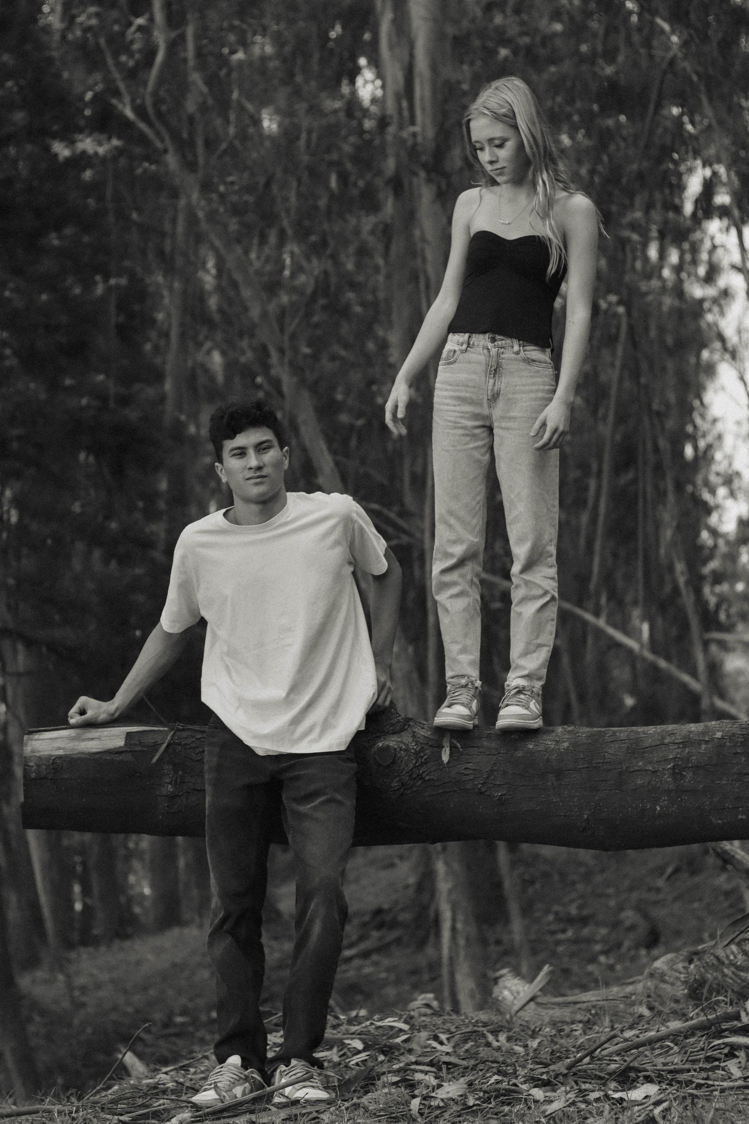 Black and white photo of teen couple in forest, girl standing on fallen log.