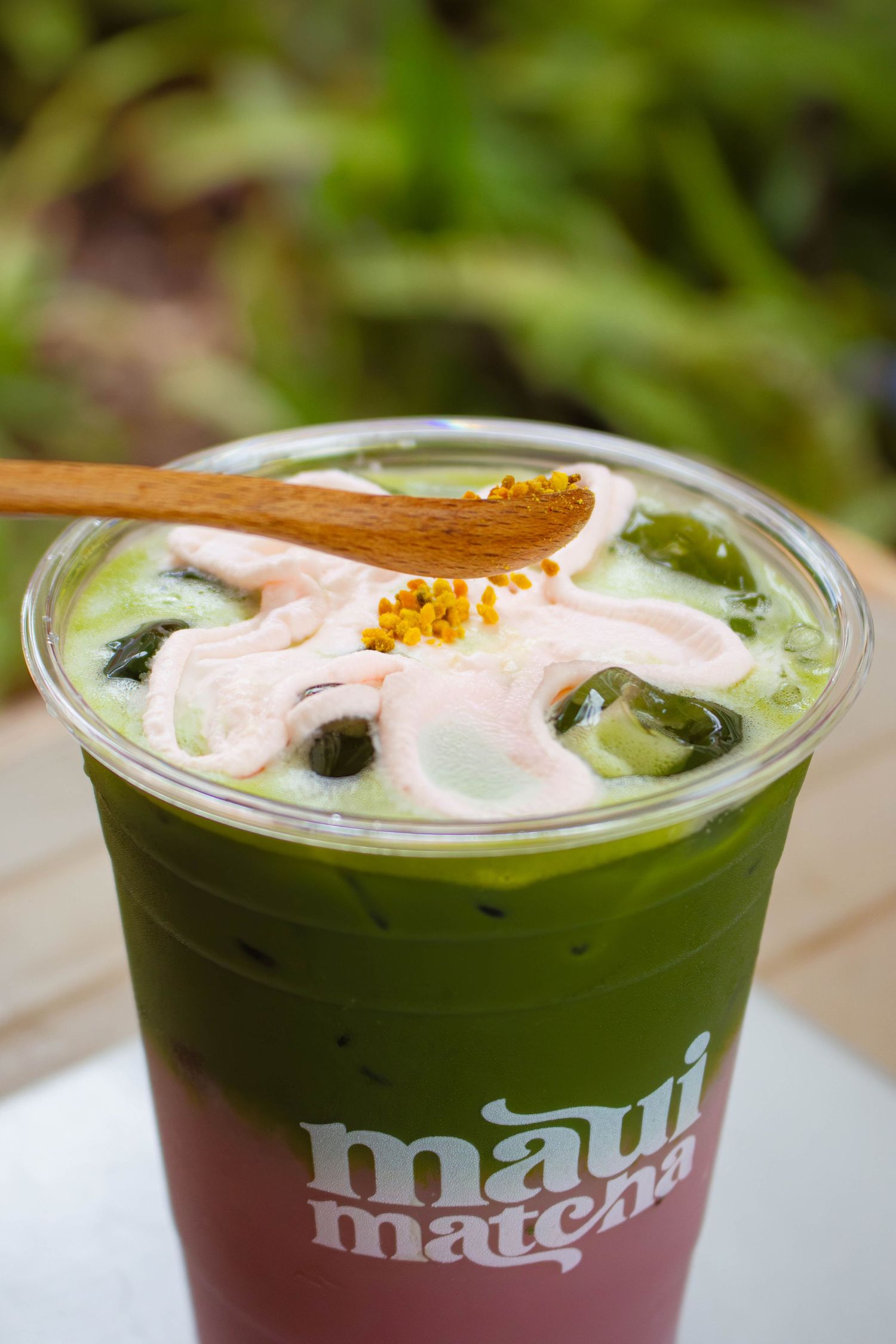 Iced matcha drink topped with pink cream and bee pollen in a Maui Matcha cup with a wooden spoon.