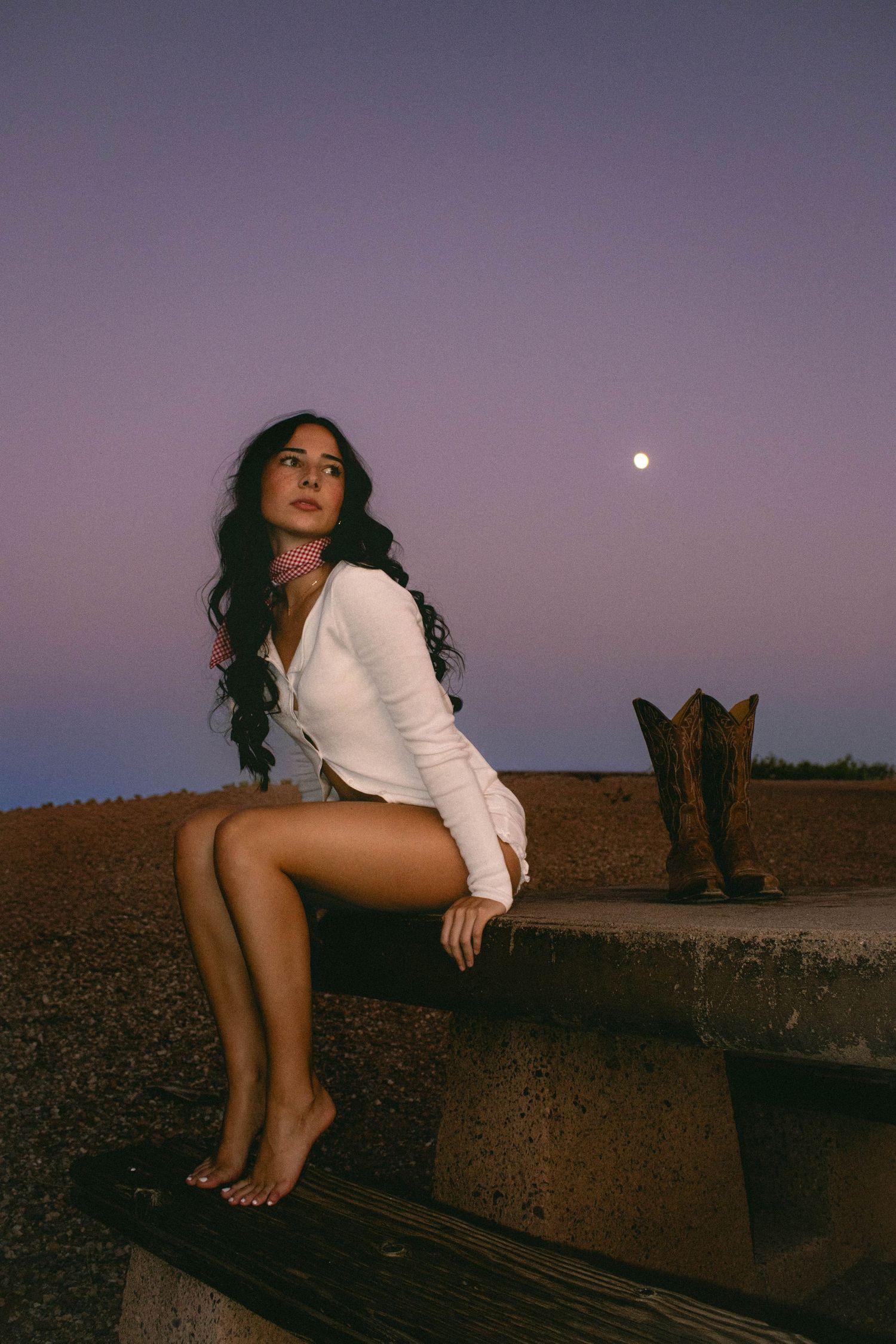 Woman in white sitting barefoot on concrete ledge under full moon at twilight.