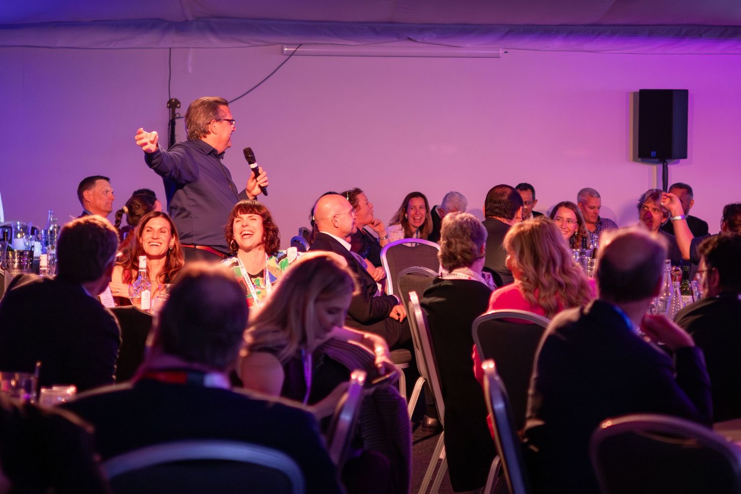 A speaker holds a microphone at a formal banquet event with seated guests under purple lighting.