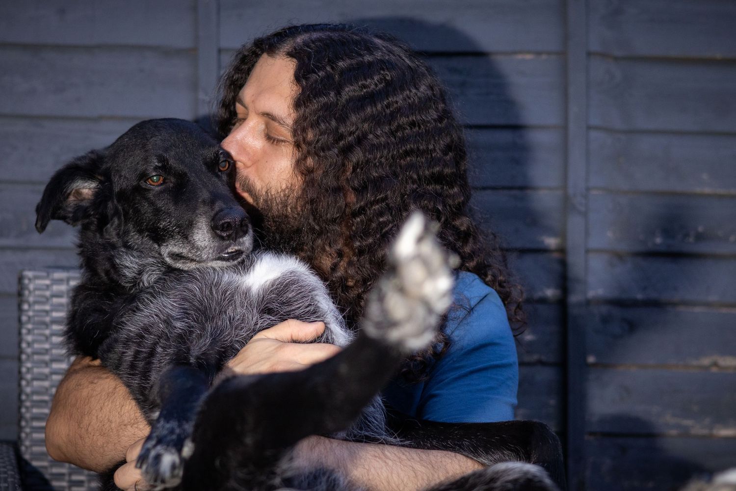 A person with long curly dark hair gently kisses a small black and white dog while seated outside.