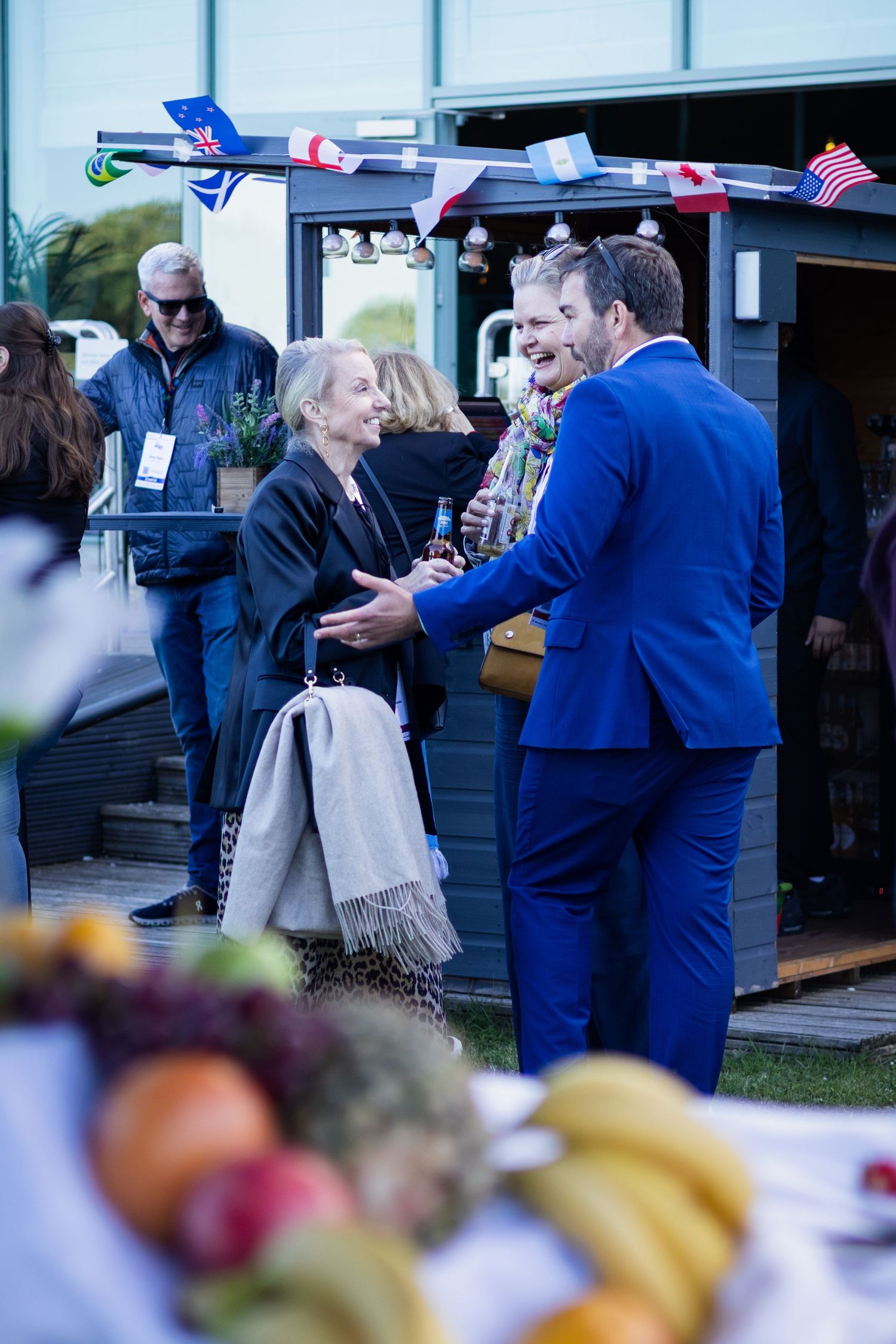 Guests networking outdoors at evening event with fruit display and international flags.