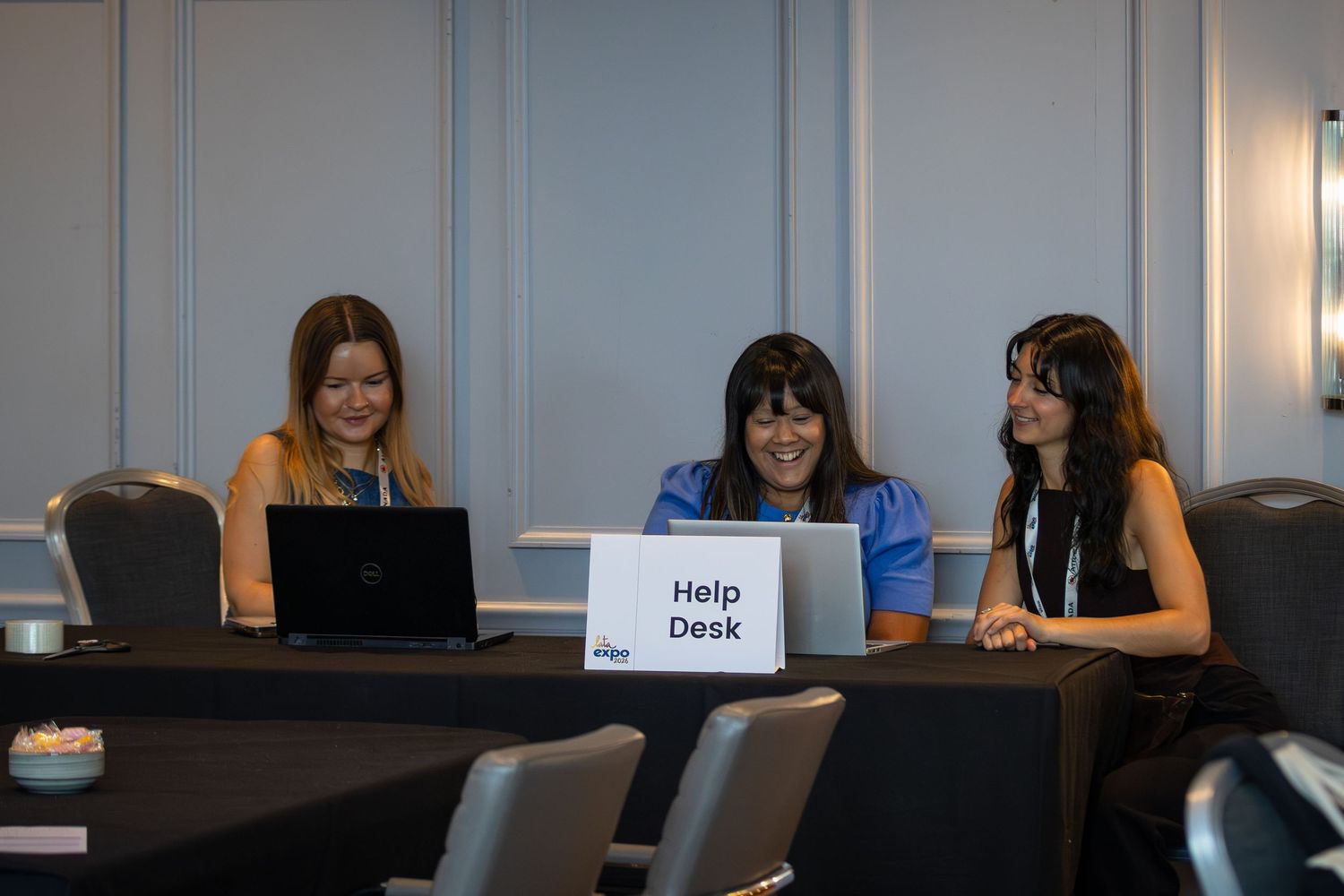 Three women smiling at a help desk with laptops at a conference event.