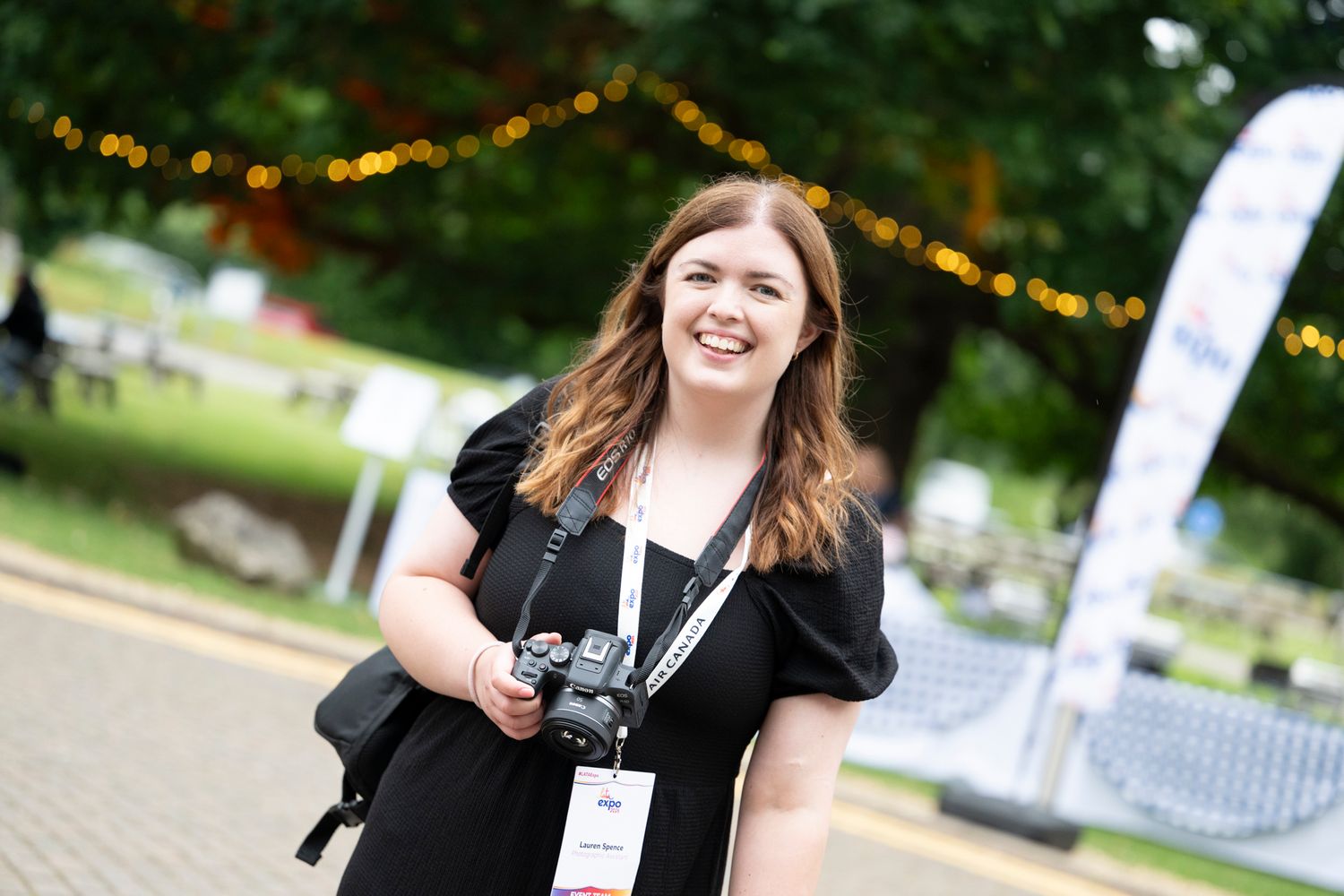 Smiling woman photographer Lauren Zagonel holding a camera at an outdoor event wearing a black dress.
