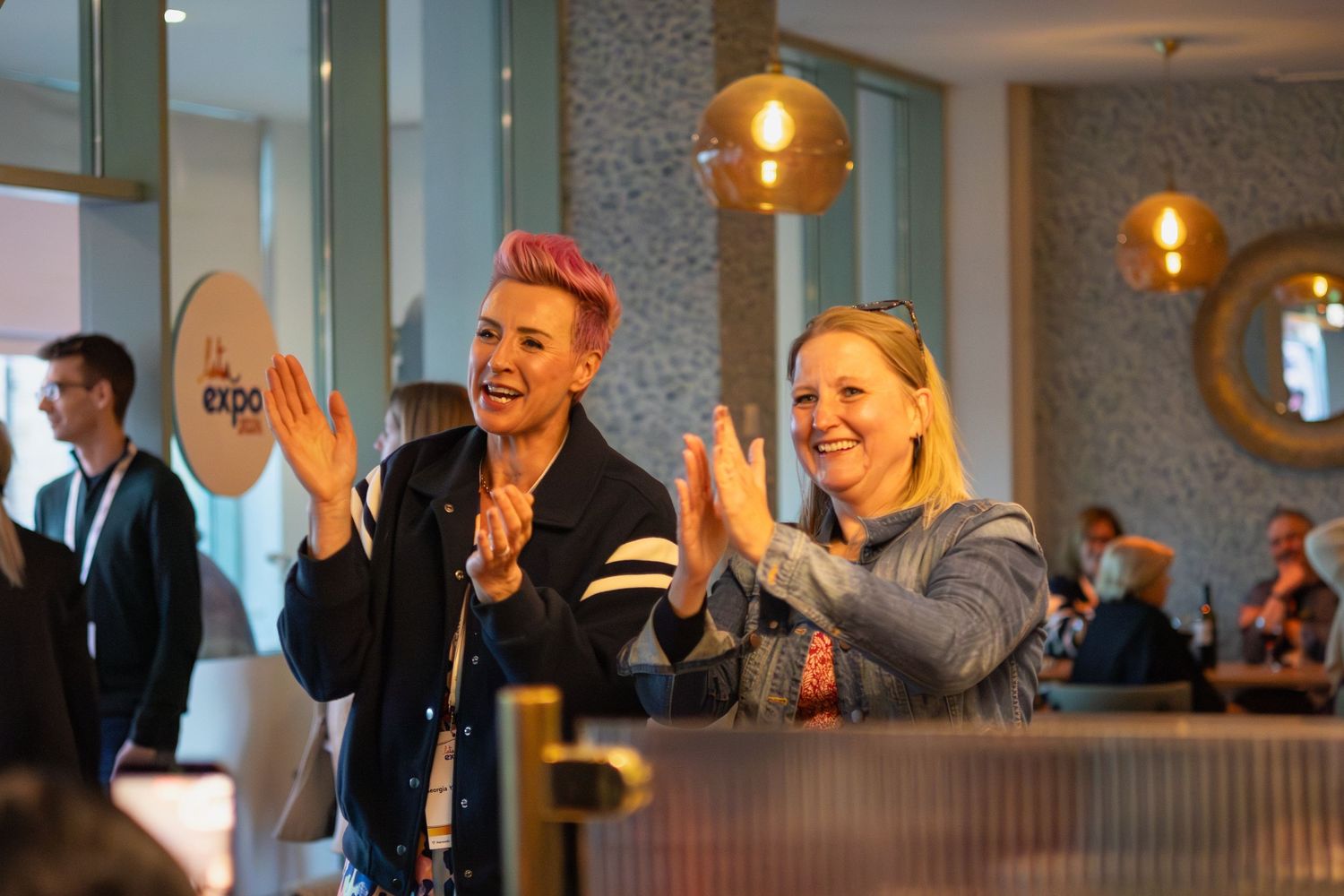Two women clapping and smiling at indoor corporate networking event with warm lighting.