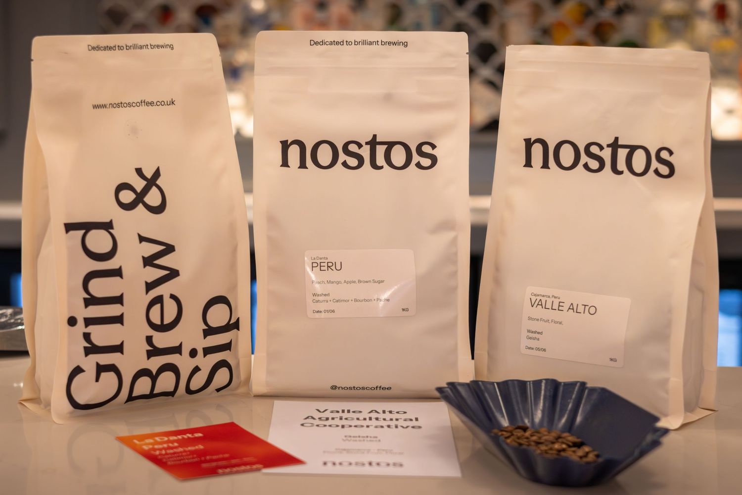 Three white Nostos coffee bags and Grind Brew Sip brands displayed on a counter.