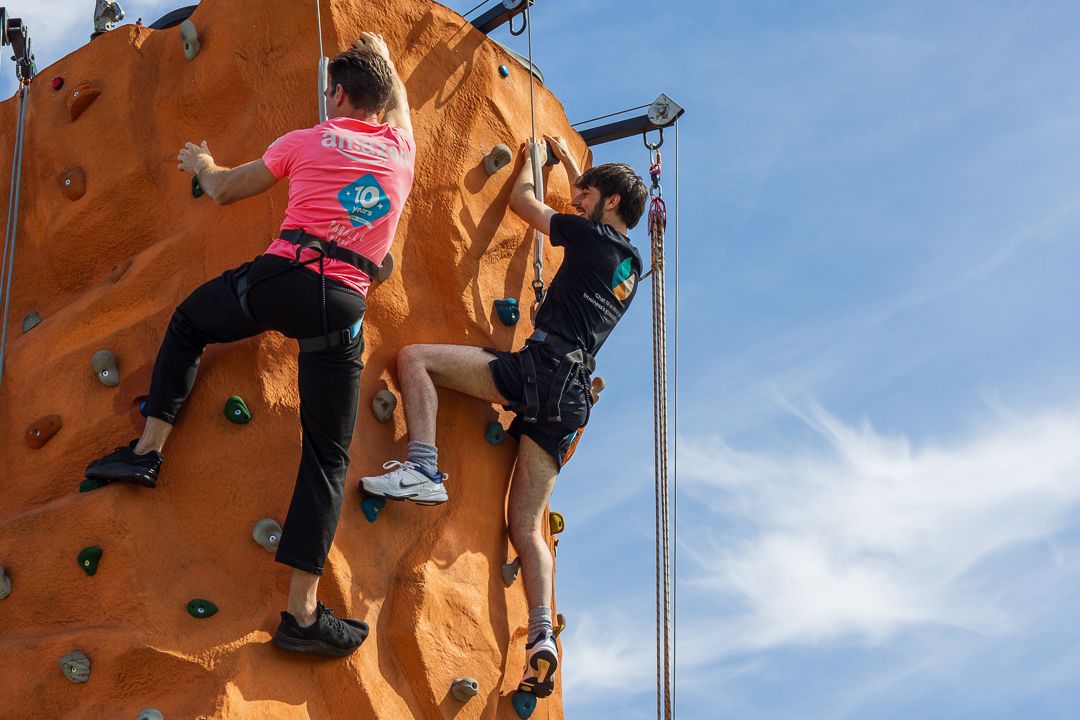 Two climbers scaling an outdoor orange rock climbing wall under a blue sky.