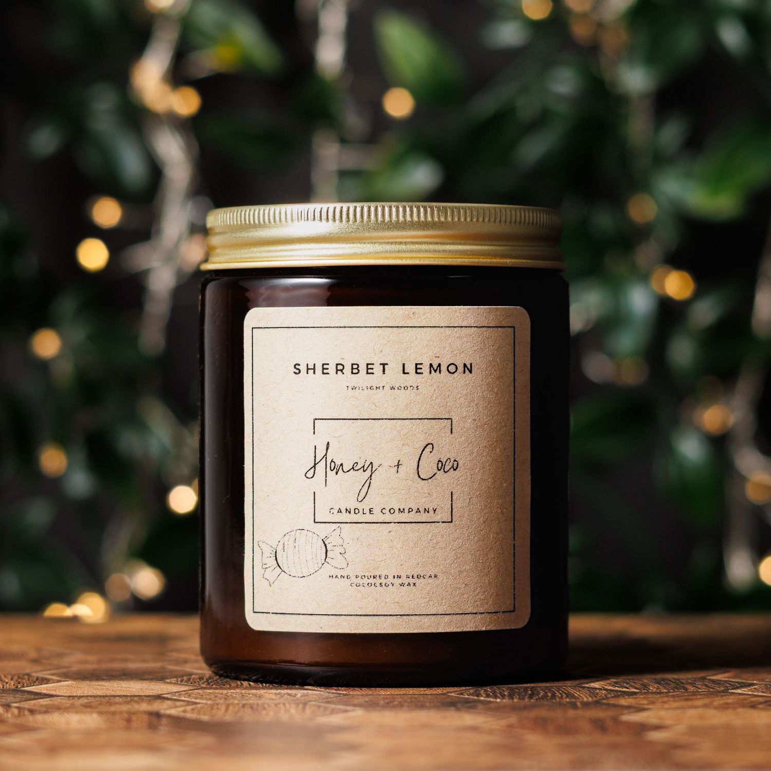 Sherbet Lemon scented candle in an amber glass jar with gold lid and Honey + Coco Candle Company label on a wooden table with fairy lights.