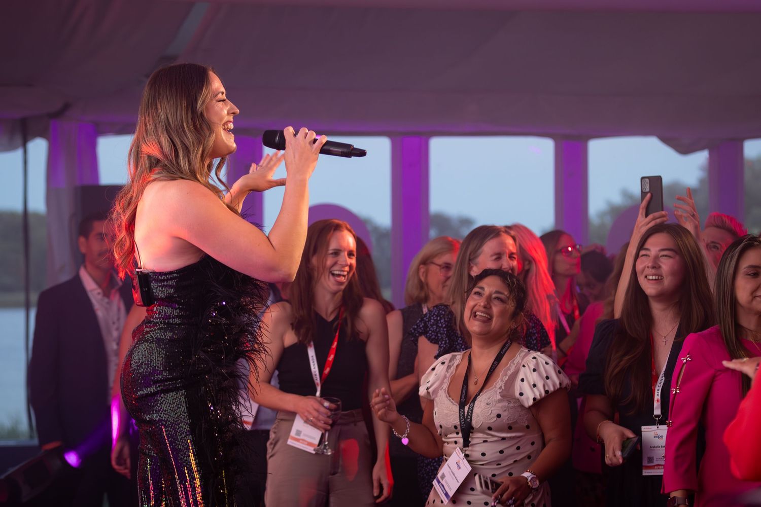 Female singer performing with microphone to an engaged crowd at an indoor event under pink lights.