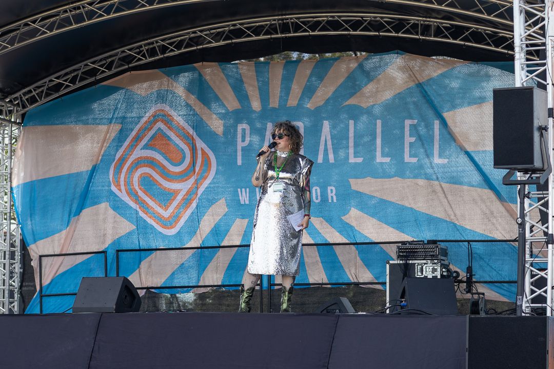 Performer on stage at Parallel Windsor festival, standing at microphone under colorful sunburst backdrop.
