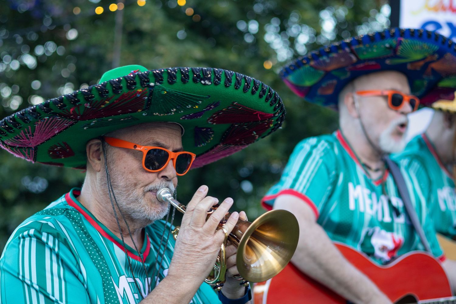Two men in sombreros and green shirts playing trumpet and guitar at an outdoor festival.