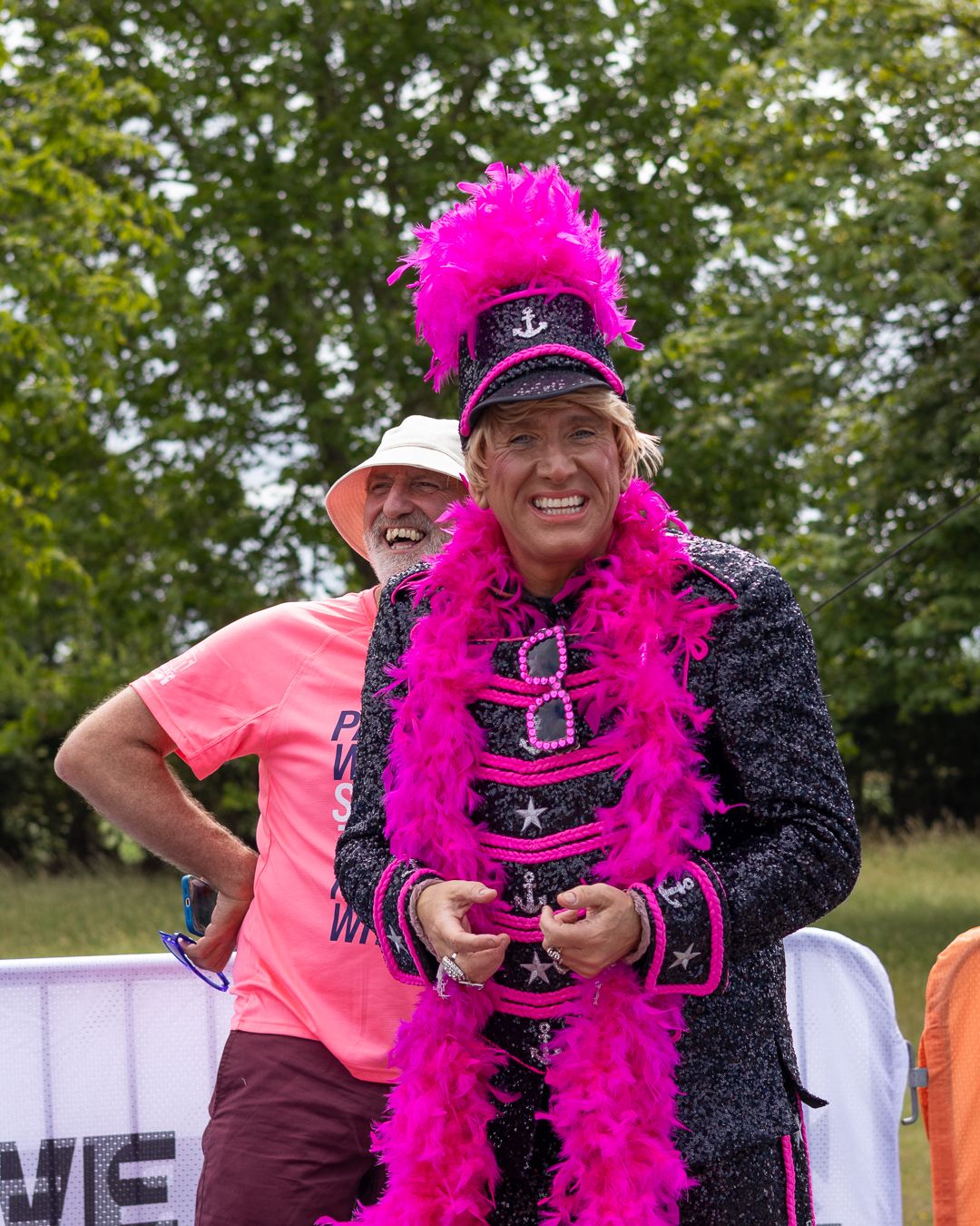 Person in vibrant pink feather boa and sequined marching band jacket with feathered hat at outdoor event.