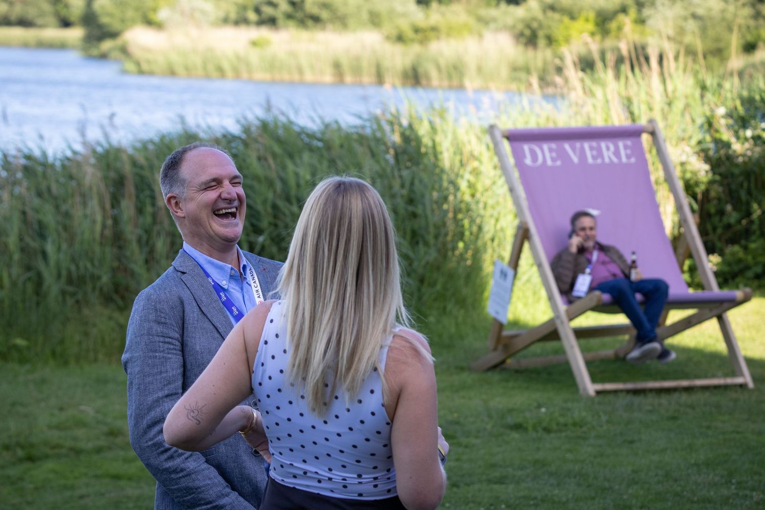 Man laughing outdoors near lake with De Vere branded deckchair and colleague in background.