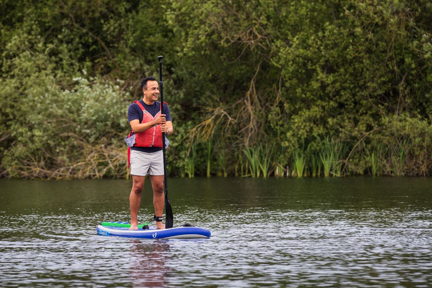 Man smiling while paddleboarding on a calm river surrounded by green trees.