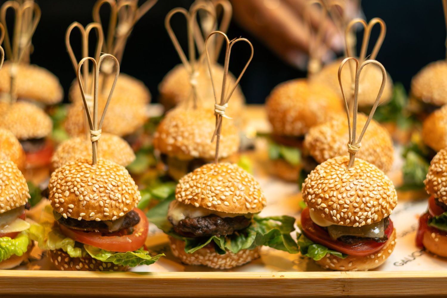 Rows of mini sesame seed bun sliders with lettuce, tomato, and meat patties displayed on a wooden tray.