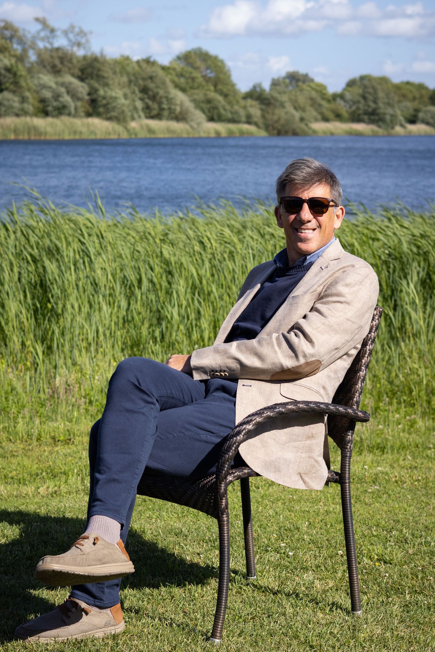 Smiling man in sunglasses and blazer relaxing on chair by scenic lakeside on sunny day.