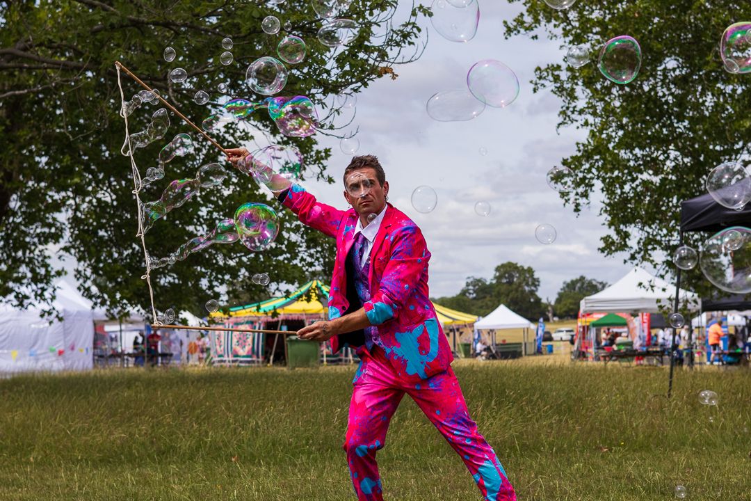 Man in bright pink suit performing bubble art at an outdoor festival on a green lawn.