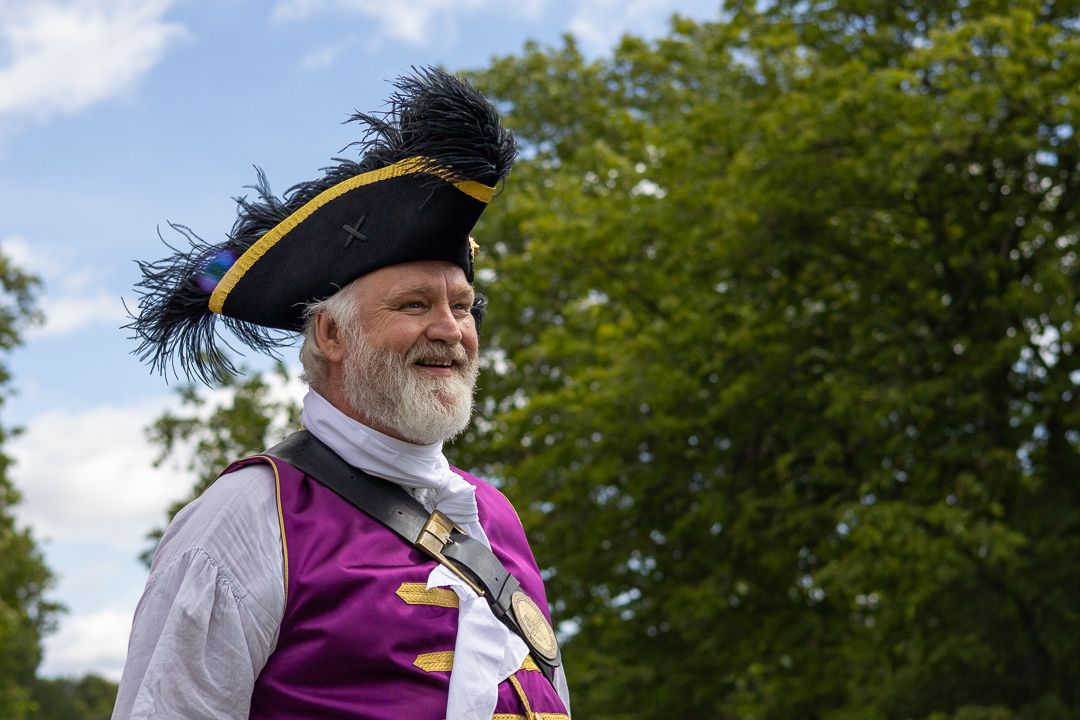Smiling bearded man in purple town crier costume with tricorn hat and feathers outdoors.