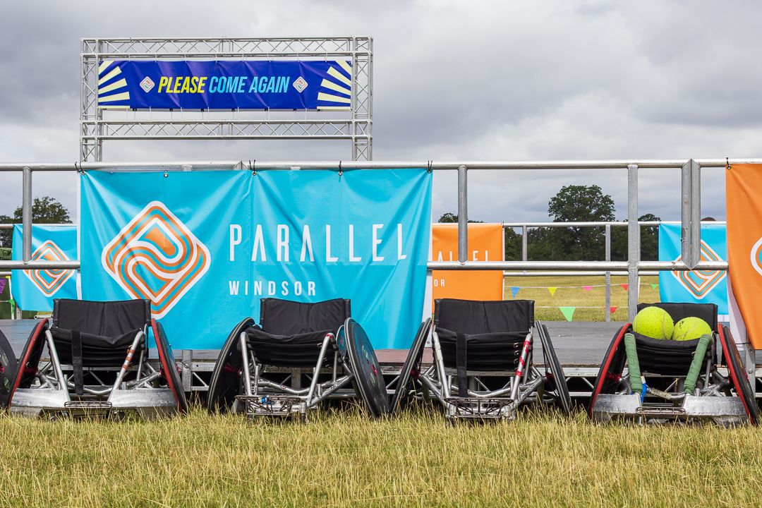Parallel Windsor wheelchair racing chairs lined up on grass at an outdoor event with a Please Come Again banner.