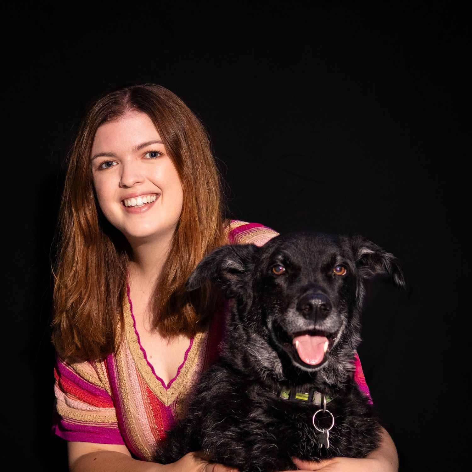 A smiling young woman with auburn hair poses with a fluffy black dog against a dark studio background.
