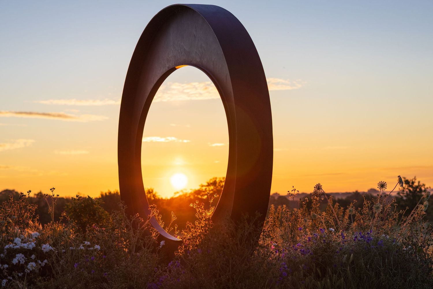 A circular metal sculpture framing a golden sunset over a wildflower meadow at dusk.