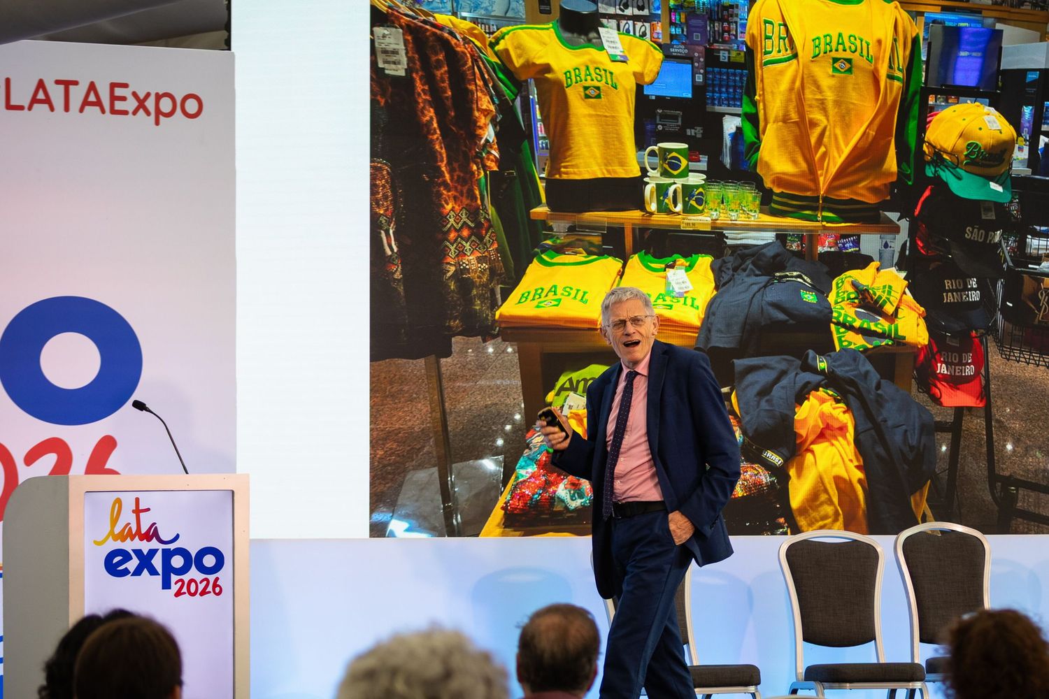 A speaker presents at LATA Expo 2024 podium with Brazilian merchandise display projected on screen behind him.