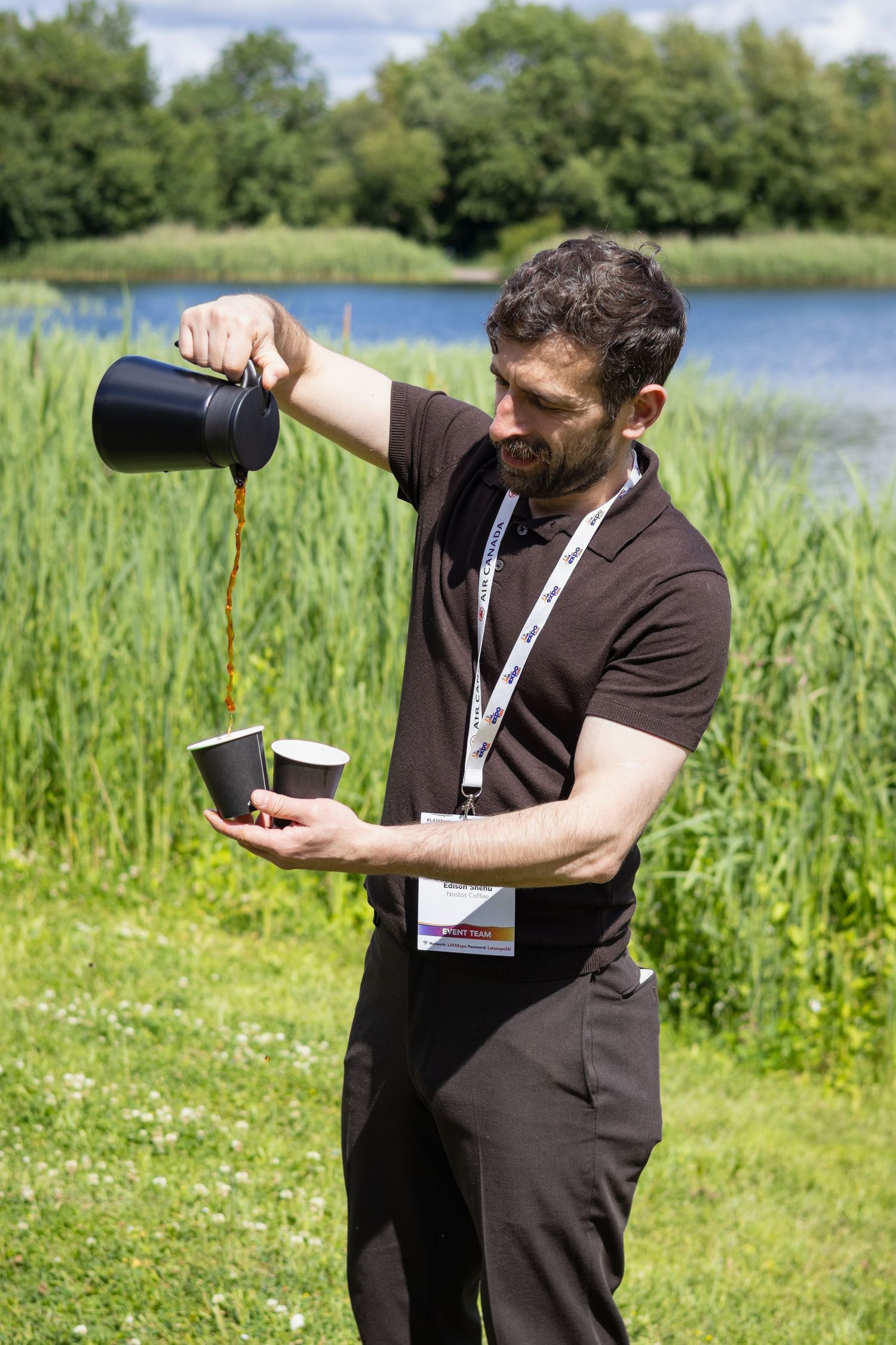 Bearded man pouring coffee into cups outdoors at lakeside corporate conference event.