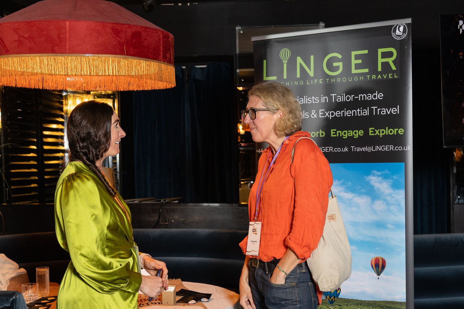 Two women networking at a Linger luxury travel event, standing near a promotional banner indoors.