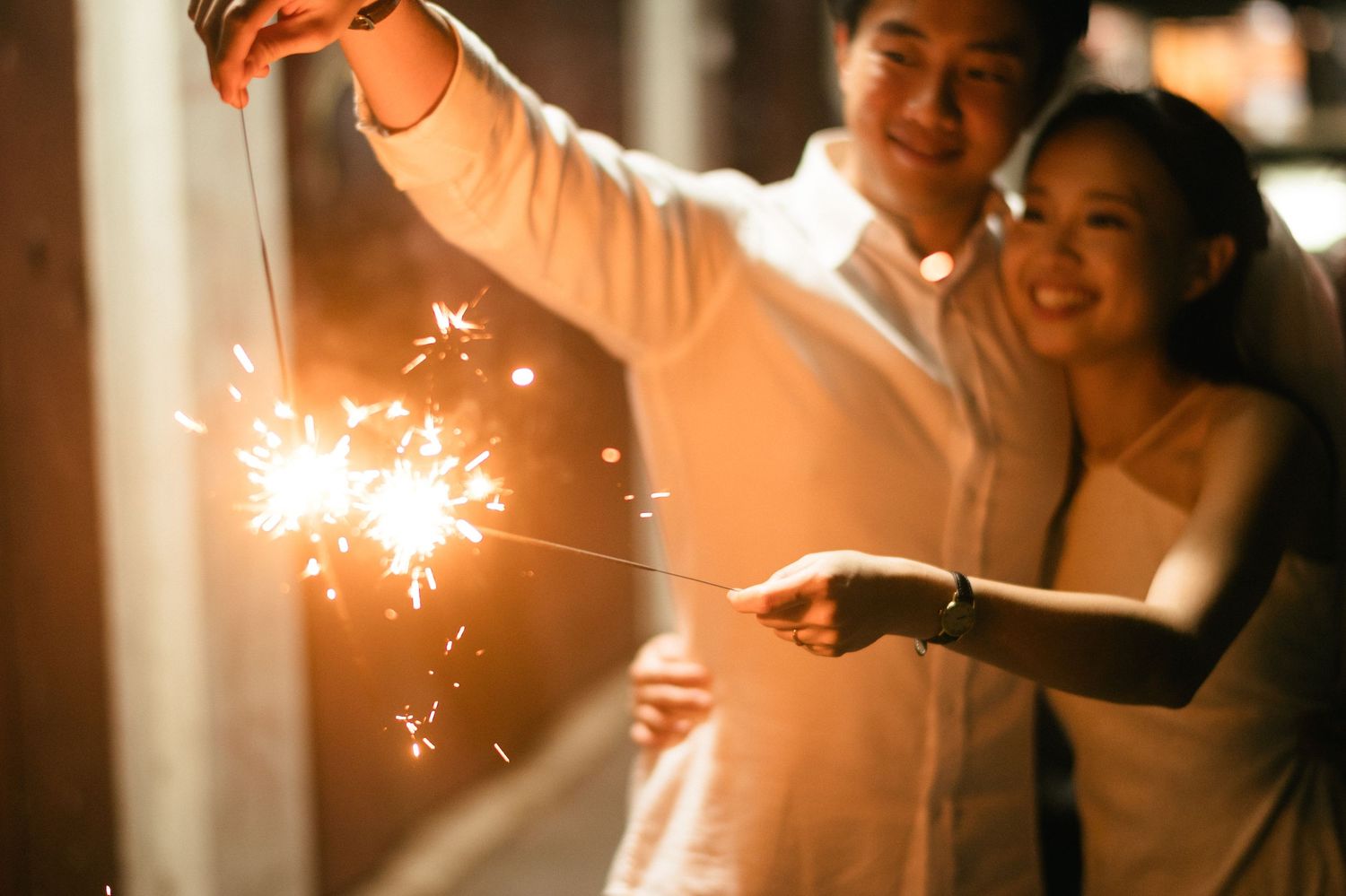 Couple holding sparklers together, smiling and celebrating at a festive event in warm golden light.
