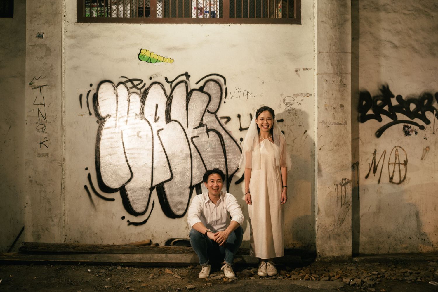 Couple posing against a graffiti-covered wall at night, man crouching and woman standing in a white dress.