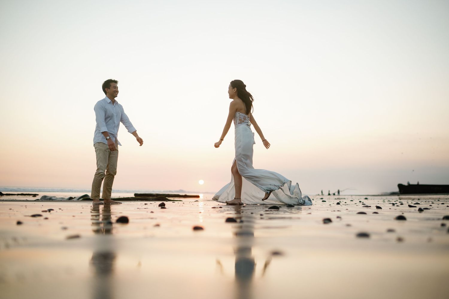 Bride in white gown and groom walking toward each other on a reflective beach at sunset.