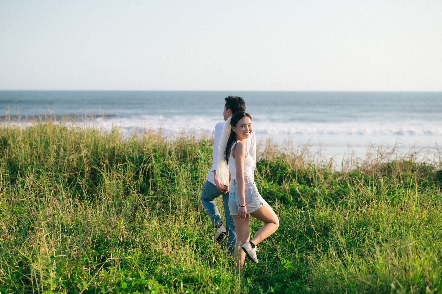 Couple walking through lush green coastal grass with ocean waves in the background on a sunny day.
