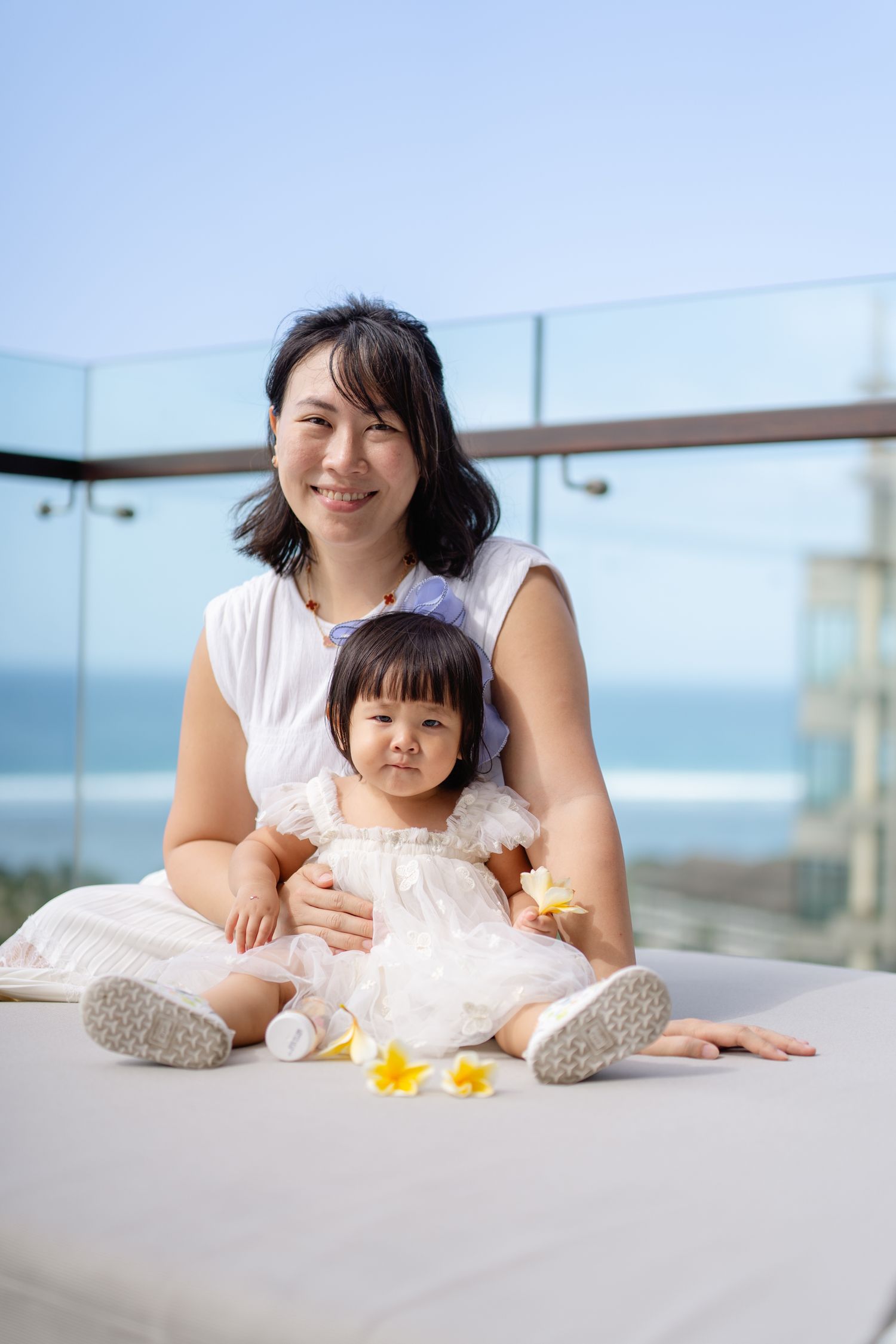 Asian mother and toddler daughter in white dresses sitting on balcony with ocean view and plumeria flowers.