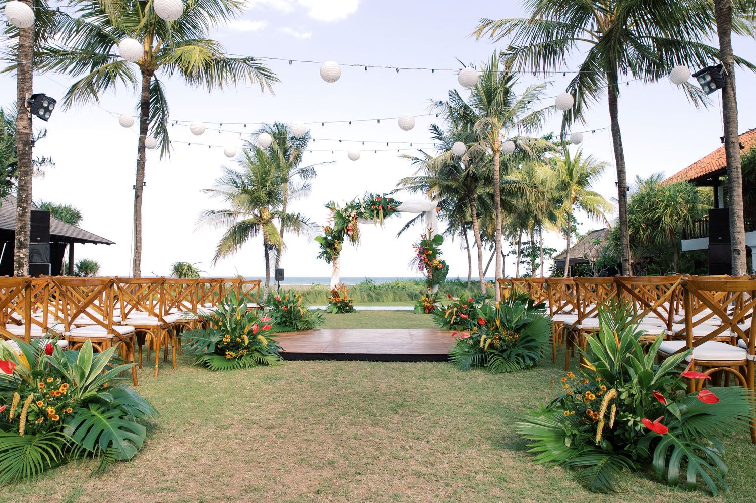 Tropical outdoor wedding ceremony setup with palm trees, string lights, wooden chairs, and lush greenery near a beach.