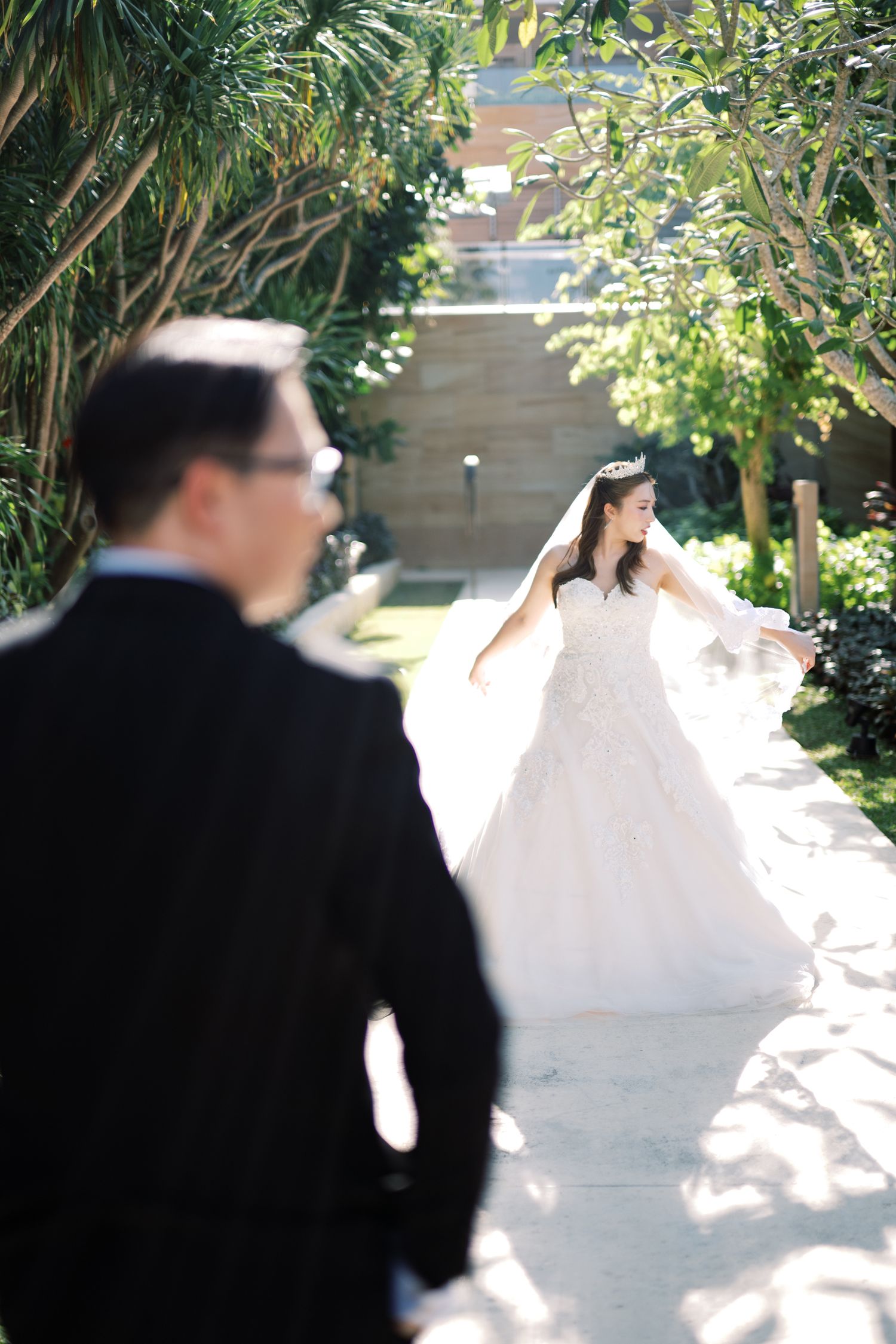 Bride in white ball gown walking toward groom in black tuxedo along a sunlit garden pathway.