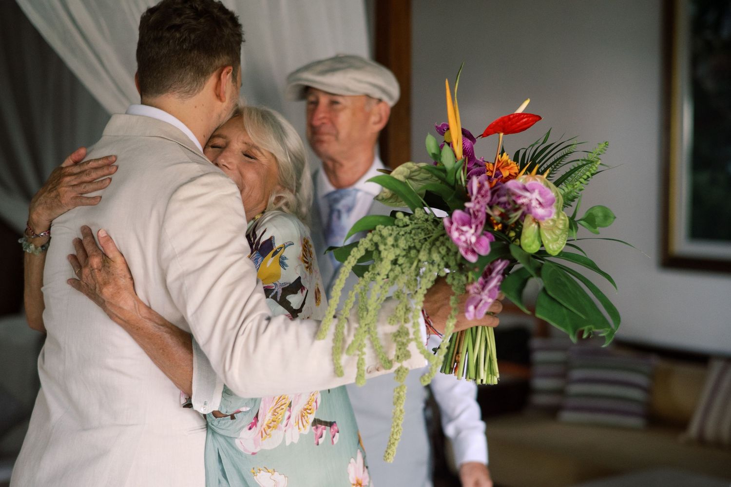 A couple embraces at a wedding while a man in a flat cap watches, with a colorful tropical bouquet in the foreground.