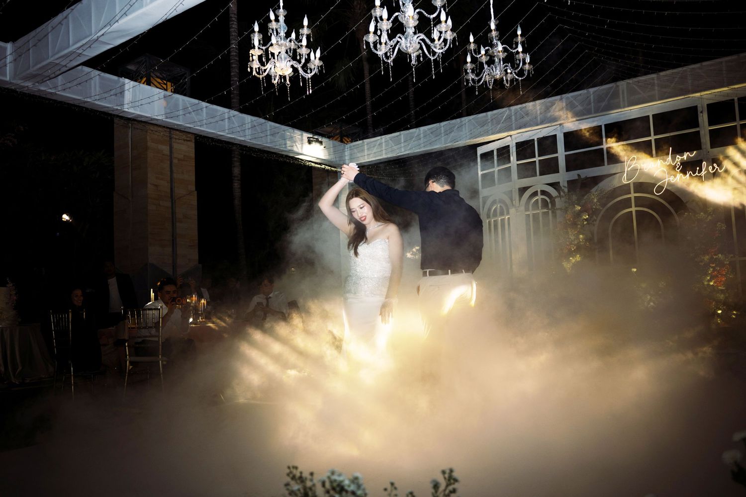 Bride and groom share first dance surrounded by dramatic fog and glowing chandeliers at elegant wedding reception.