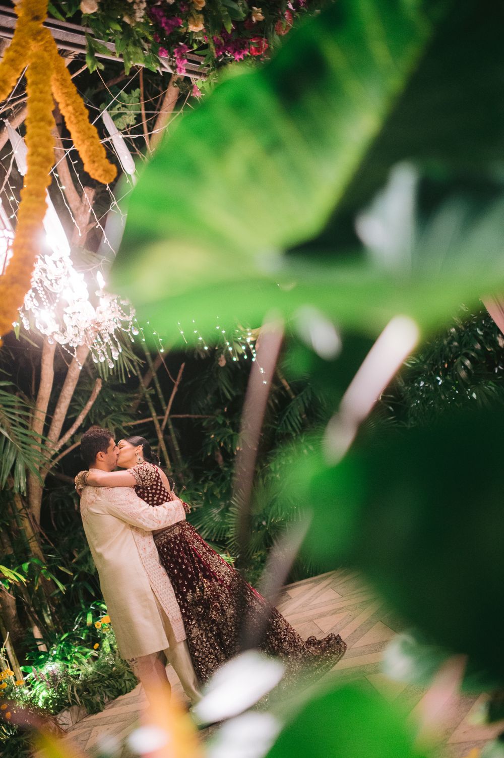 A couple embraces romantically amid lush tropical greenery and twinkling lights at an outdoor Indian wedding.