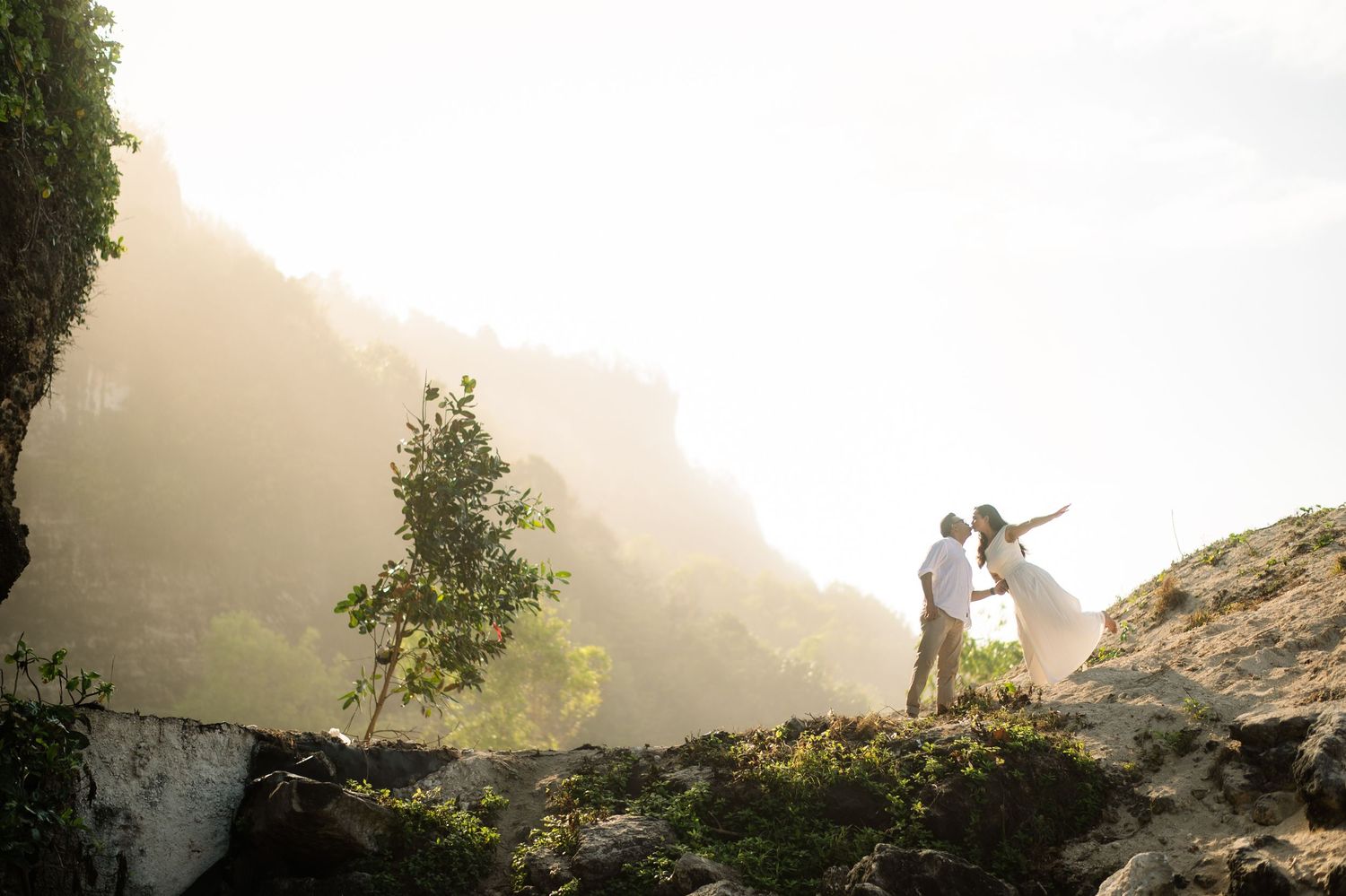 Couple in wedding attire standing on rocky cliffside with misty mountains and lush greenery in background.