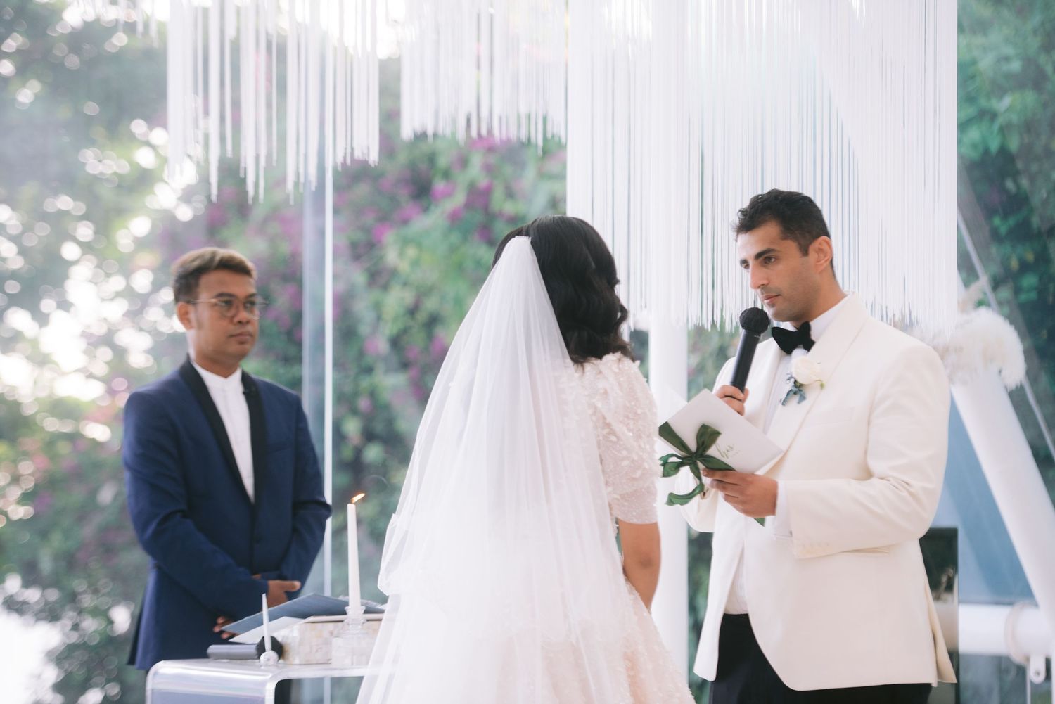 Groom reads vows to bride in white dress and veil at elegant outdoor wedding ceremony with white floral backdrop.