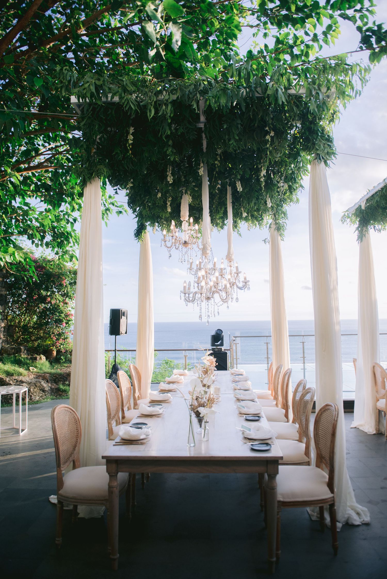Elegant outdoor wedding reception with long dining table, crystal chandelier, white drapes, and ocean view.