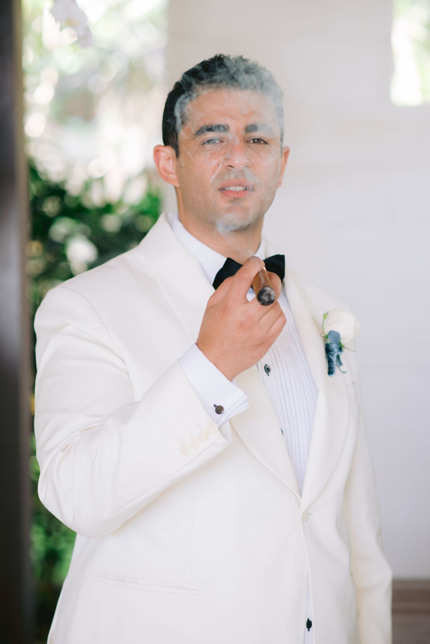 Groom in white tuxedo jacket and black bow tie smoking a cigar with a white boutonniere at a wedding.
