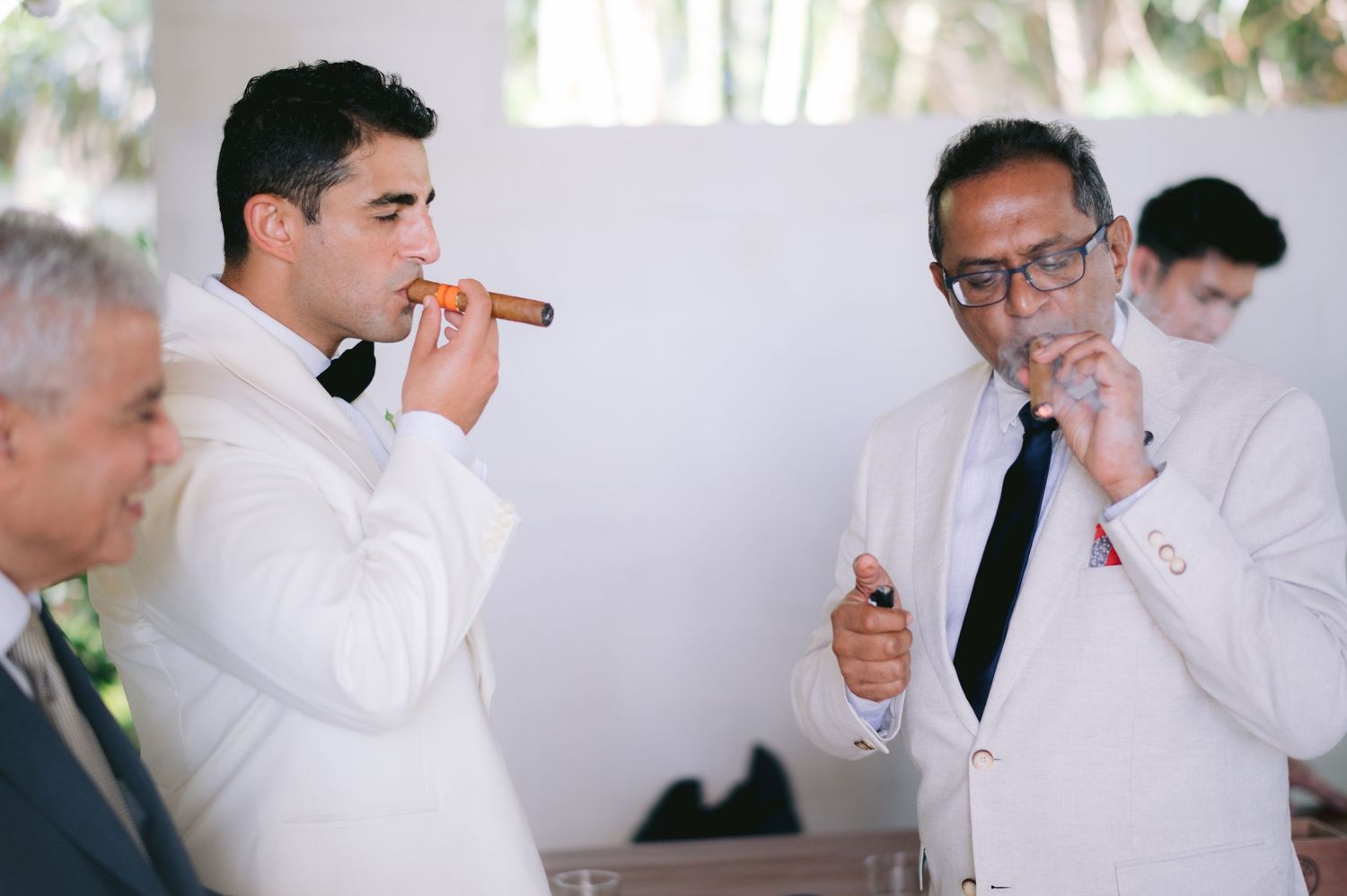 Two men in white suits smoking cigars at a formal event, with a third man visible in the background.