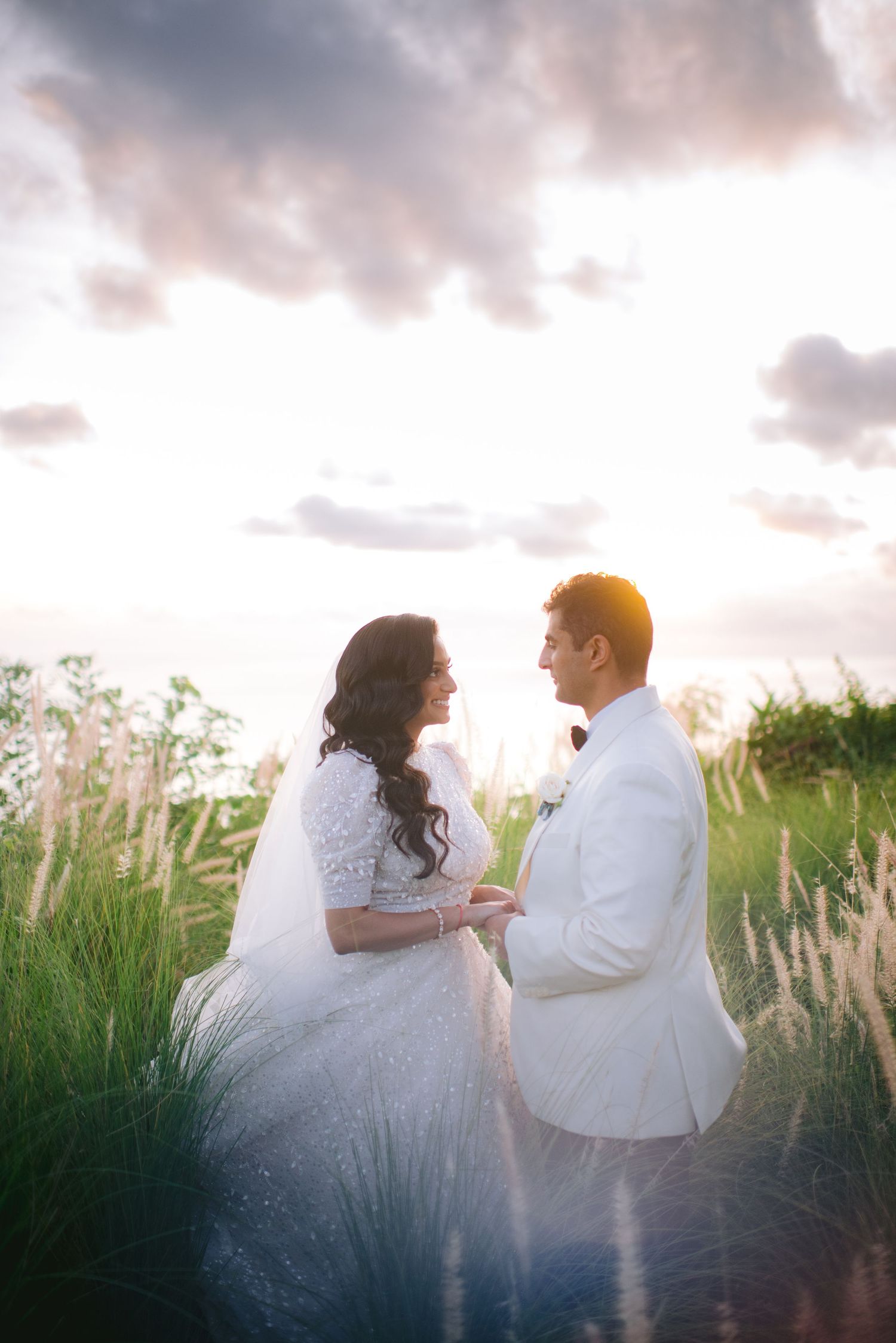 Bride and groom share a romantic moment in a sunlit field, wearing white wedding attire with dramatic cloudy sky backdrop.