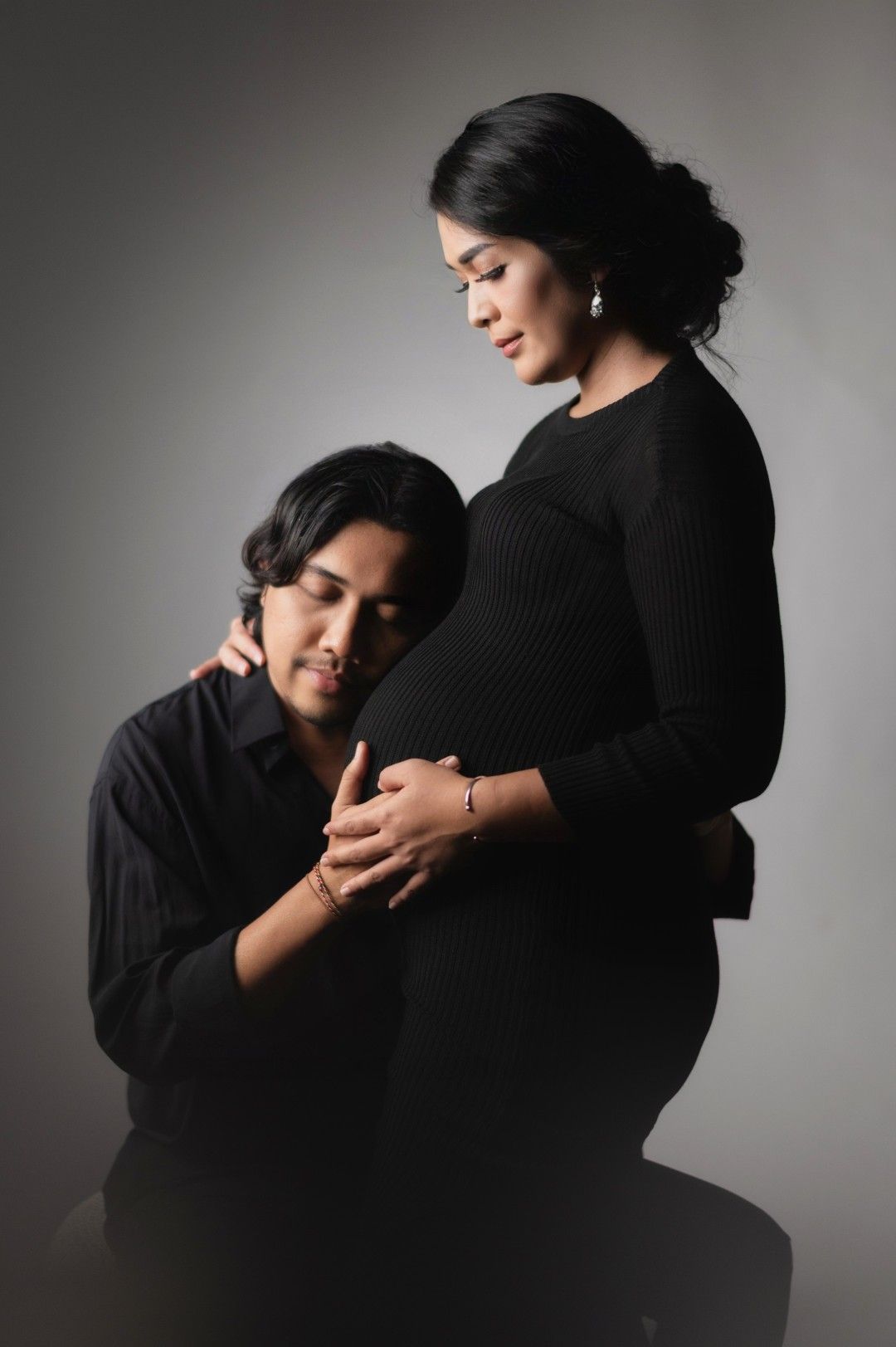 Couple in black outfits posing for maternity photo, man kneeling beside pregnant woman against gray background.