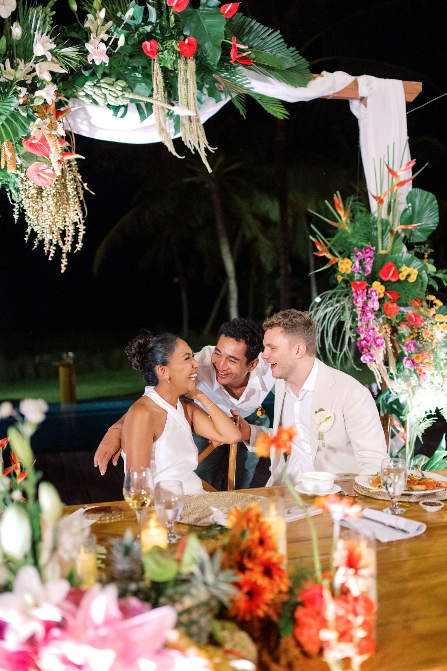 Bride and groom laughing with guest at tropical wedding reception under lush floral arch with colorful blooms at night.