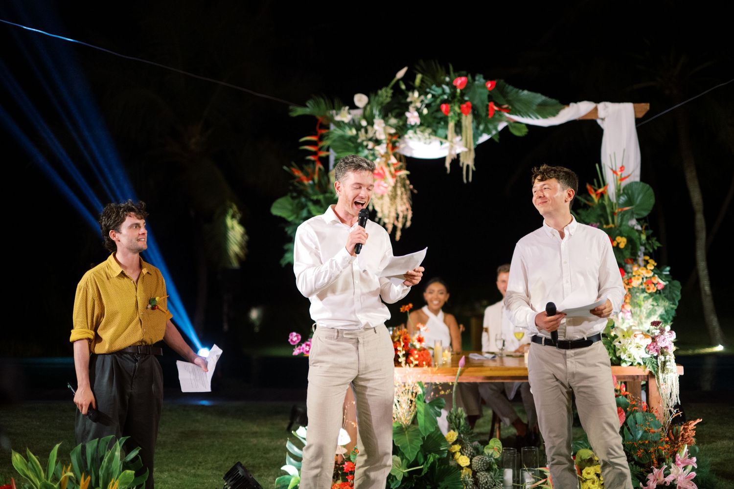 Three men deliver heartfelt wedding speeches at night under string lights before floral tropical ceremony arch.